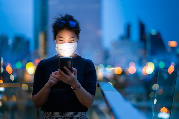 Atrakcyjna azjatycka kobieta nosi maskę chroniącą rękę używa technologii smartfona komunikacja z tłem bokeh lekki budynek miejski koncepcja pomysłów biznesowych w tle