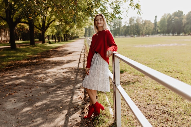 Atrakcyjna blondynka pozuje w stylowym czerwonym stroju w jesiennym parku. Ładna dziewczyna na sobie białą sukienkę o odpowiednim czasie na świeżym powietrzu.