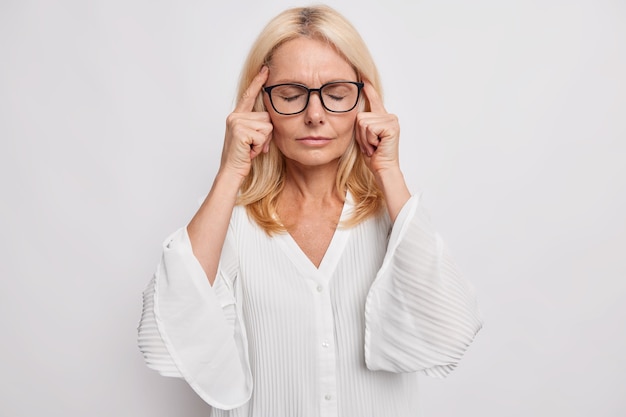 Atrakcyjna Blondynka W średnim Wieku Trzyma Palce Na Skroniach Cierpi Na Ból Głowy Próbuje Sobie Przypomnieć Coś Ważnego Stoi Z Zamkniętymi Oczami Nosi Okulary Biała Bluzka Stoi Przed Trudnym Problemem