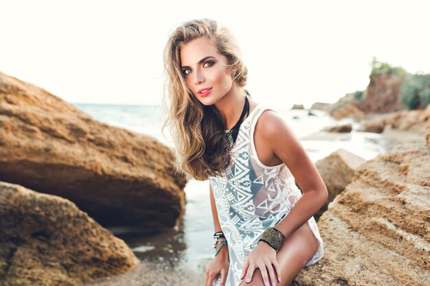 Atrakcyjna blondynka z długimi włosami siedzi na kamieniu na kamienistej plaży na tle zachodu słońca.