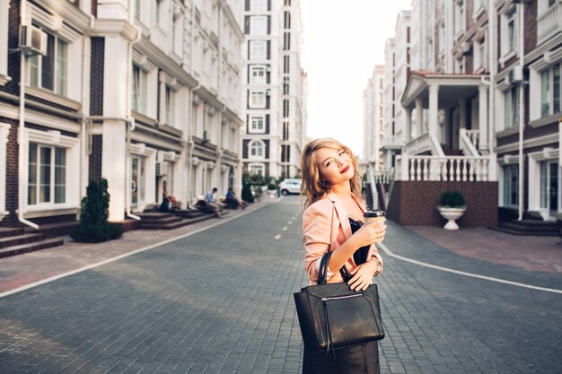 Atrakcyjna blondynka z winnymi ustami, chodzenie z filiżanką kawy w koralowej kurtce na ulicy. Nosi czarną torbę