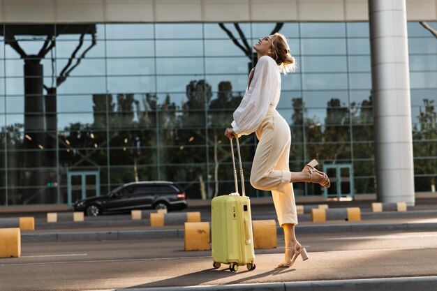 Atrakcyjna blondynka zalotnie unosi nogę i pozuje z bagażem Szczęśliwa piękna dziewczyna w białej bluzce i beżowych spodniach trzyma walizkę w pobliżu lotniska