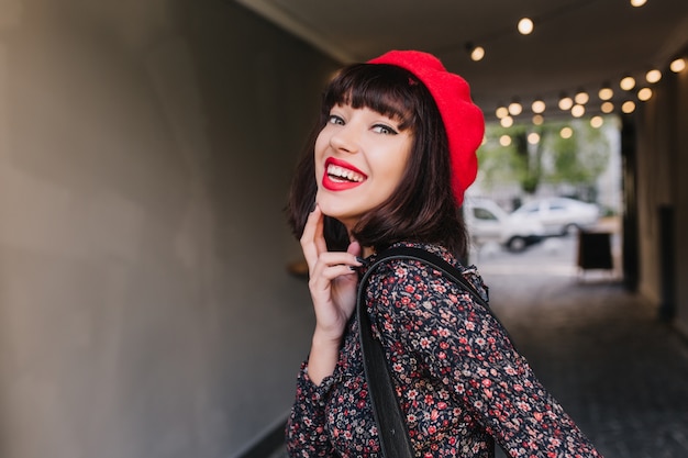 Atrakcyjna brunetka dziewczyna w stylowy strój vintage zalotnie pozuje ze szczerym uśmiechem dotykając jej podbródka. Szczegół portret eleganckiej młodej kobiety w modnych francuskich ubraniach, śmiejąc się i zabawy