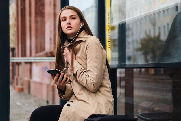 Atrakcyjna dorywcza dziewczyna w słuchawkach z telefonem komórkowym w zamyśleniu czekająca na transport publiczny na przystanku autobusowym na świeżym powietrzu