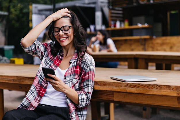 Atrakcyjna dziewczyna bawi się włosami podczas czytania wiadomości telefonicznej. Zewnątrz zdjęcie emocjonalnej studentki stwarzających z smartphone i laptopa.