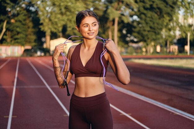 Atrakcyjna dziewczyna fitness w stylowej odzieży sportowej z skakanką na szyi marzycielsko trenująca na stadionie miejskim