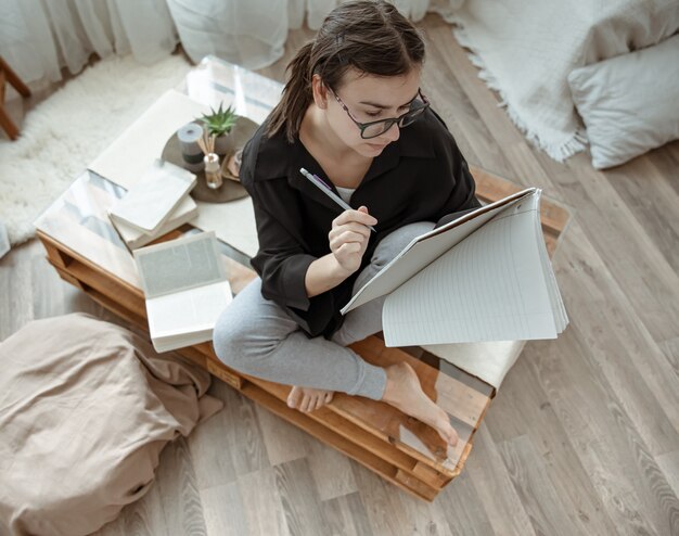 Atrakcyjna dziewczyna w domu siedzi z notatnikiem i piórem wśród widok z góry książek.