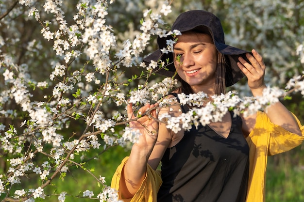 Atrakcyjna dziewczyna w kapeluszu wśród kwitnących drzew cieszy się zapachem wiosennych kwiatów