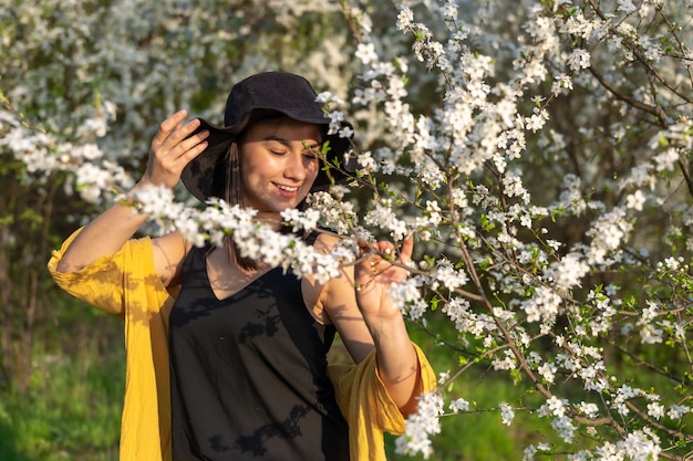 Atrakcyjna dziewczyna w kapeluszu wśród kwitnących drzew cieszy się zapachem wiosennych kwiatów