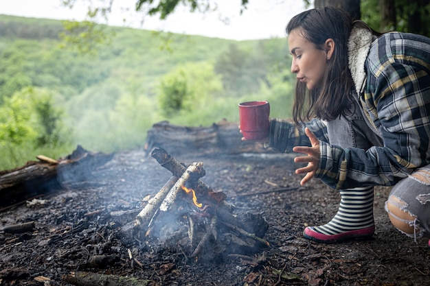 Atrakcyjna dziewczyna z filiżanką w dłoni rozgrzewa się przy ognisku w lesie.