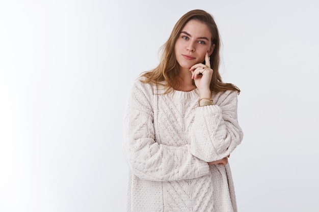 Atrakcyjna inteligentna delikatna młoda europejska kobieta ubrana w luźny przytulny ciepły sweter odpoczynek przy kominku przechylająca głowę dotykająca policzka ciesząca się komfortem uśmiechnięta szczęśliwie, zrelaksowana beztroska białe tło