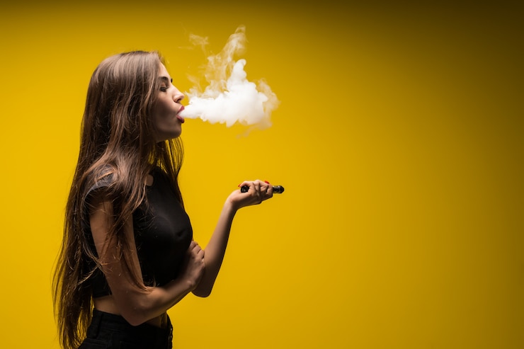 Bezpłatny plik Zdjęcie atrakcyjna kobieta stojąca i vaping na żółtej ścianie.