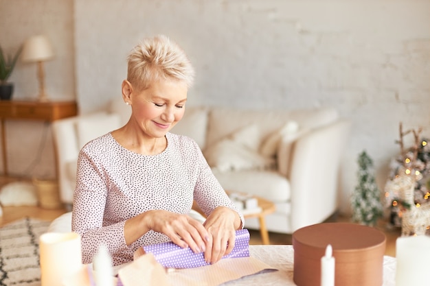 Atrakcyjna kobieta w średnim wieku z krótką blond fryzurą siedzi przy stole w salonie, pakując prezent dla męża w papier prezentowy