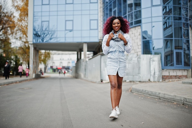 Atrakcyjna, kręcona afroamerykanka w dżinsowej sukience postawiona na tle nowoczesnego wielopiętrowego budynku