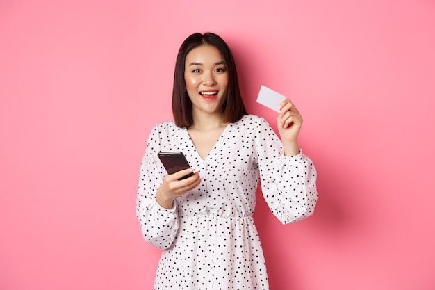 Atrakcyjna młoda Azjatka zamawia online, trzymając kartę kredytową i telefon komórkowy, robiąc zakupy przez internet...