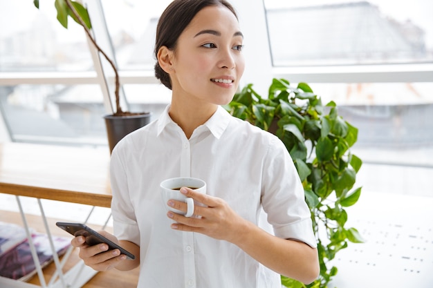 Atrakcyjna młoda azjatycka bizneswoman korzystająca z telefonu komórkowego podczas przerwy na kawę w kawiarni w pomieszczeniu