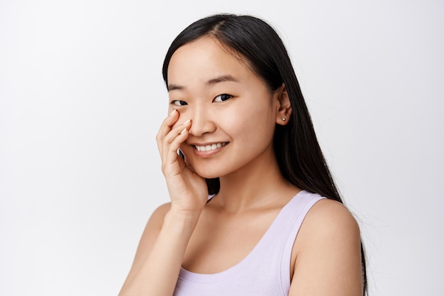 Atrakcyjna młoda azjatycka kobieta ze świecącą, zdrową skórą, uśmiechniętą i dotykającą twarzy opuszkami palców, stojącą na białym tle