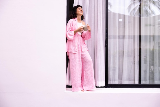 Atrakcyjna młoda brunetka kaukaska uśmiechnięta trzymająca filiżankę kawy w rękach rano stojąc przy oknie białego domu Koncepcja komfortu w domu