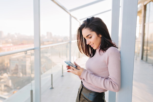 Atrakcyjna młoda kobieta brunetka pisania na telefon na tarasie z widokiem na duże miasto. Słoneczny poranek, odprężający czas, wesołe emocje, czarujący, elegancki wygląd.
