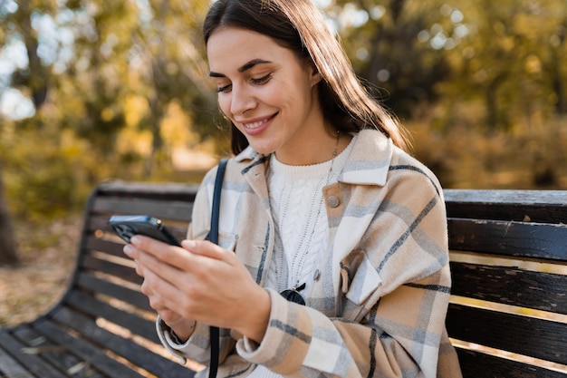 Atrakcyjna młoda kobieta chodząca jesienią ubrana w kurtkę za pomocą telefonu