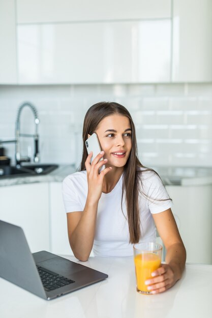 Atrakcyjna młoda kobieta opowiada na telefonie komórkowym podczas gdy stojący na kuchni z szkłem sok