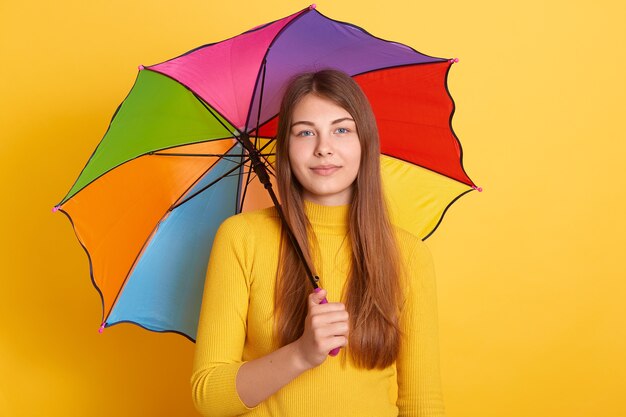 Atrakcyjna młoda kobieta, stojąca pod wielobarwnym parasolem i ubrana w żółty sweter