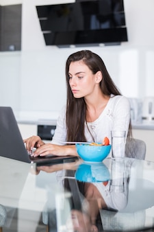 Atrakcyjna młoda kobieta używa laptop przy śniadaniem i obsiadanie w kuchni.