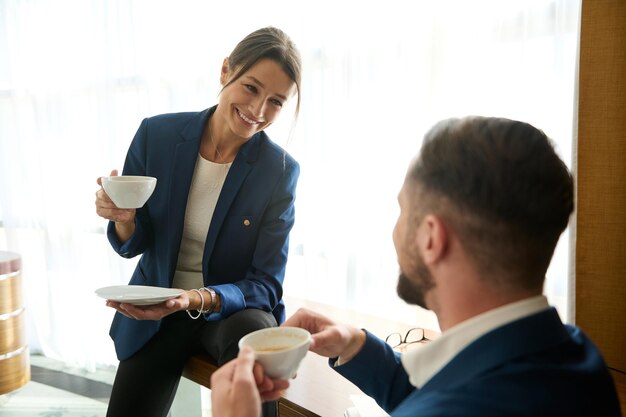 Atrakcyjna młoda kobieta w garniturze siedzi przy stole w pokoju hotelowym i uśmiecha się słodko rozmawiając ze swoim szefem lub partnerem biznesowym przy filiżance kawy. relacje partnerskie i bliskość w pracy