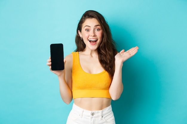 Atrakcyjna młoda kobieta w letnich ubraniach glamour, pokazująca pusty ekran smartfona, dysząca zdumiona, polecająca aplikację, stojąca na niebieskim tle.