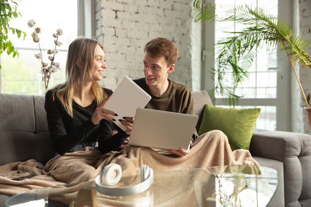 Atrakcyjna młoda para za pomocą urządzeń razem, tablet, laptop, smartfon, słuchawki bezprzewodowe. Komunikacja, koncepcja gadżetów. Technologie łączące ludzi w samoizolacji. Styl życia w domu.