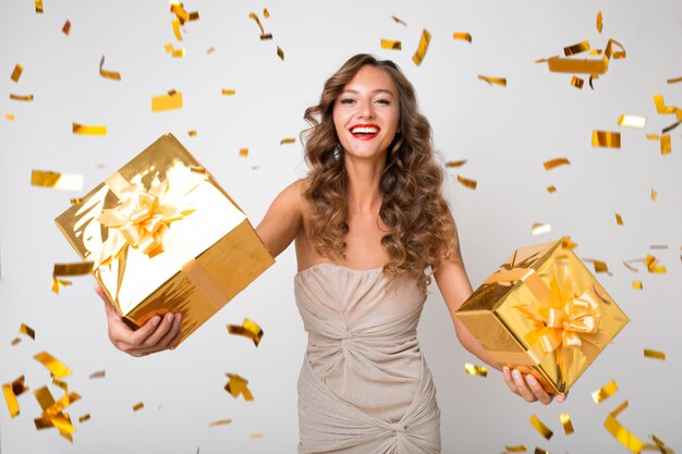 Atrakcyjna młoda stylowa kobieta obchodzi nowy rok, trzymając prezenty w pudełku, złote konfetti latające, uśmiechnięta szczęśliwa, ubrana w sukienkę