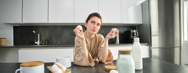 Bezpłatne zdjęcie atrakcyjna młoda, wesoła dziewczyna piecze w kuchni, wyrabiając ciasto, trzymając książkę kucharską i mając pomysły