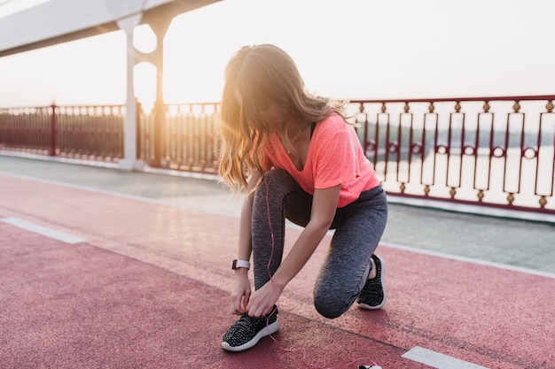 Atrakcyjna modelka w modnych ubraniach przygotowuje się do maratonu. Odkryty strzał brunetki zawiązuje sznurówki na stadionie.