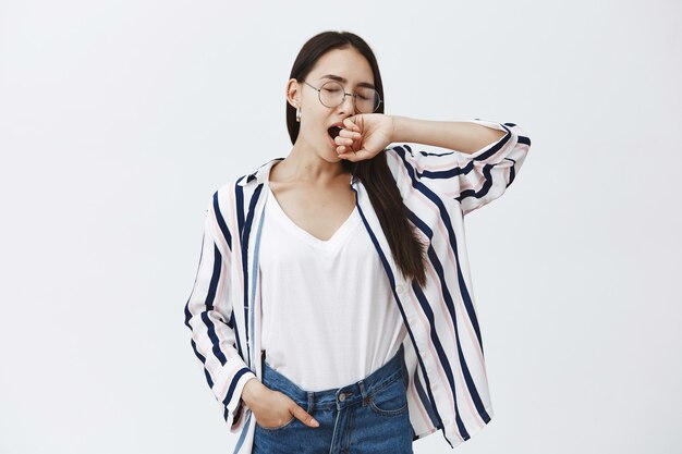 Atrakcyjna modna blogerka w pasiastej bluzce i okularach, rozciągająca się z zamkniętymi oczami i ziewająca, zakrywająca usta ramieniem, zmęczona i senna, chcąca wyjść z domu