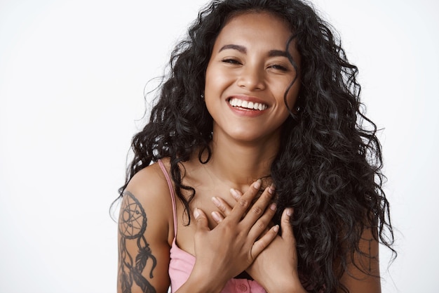 Atrakcyjna nowoczesna kobieta z kręconymi włosami z tatuażami, trzymająca ręce na klatce piersiowej wdzięczna i wzruszona, śmiejąca się i uśmiechnięta, ciesząca się dotykaniem uroczej randki, białej ściany