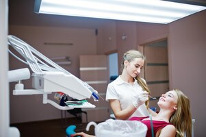Bezpłatne zdjęcie atrakcyjna pacjentka w czerwonofioletowej sukience leżąca na fotelu dentystycznym, podczas gdy dentystka leczy zęby specjalnymi narzędziami