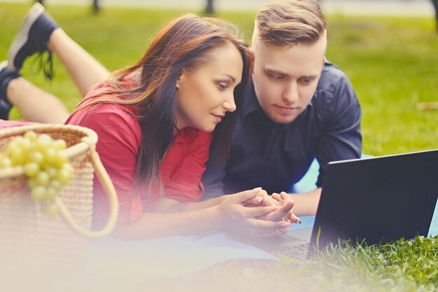 Atrakcyjna para leży na kocu na trawniku i korzysta z laptopa na pikniku.