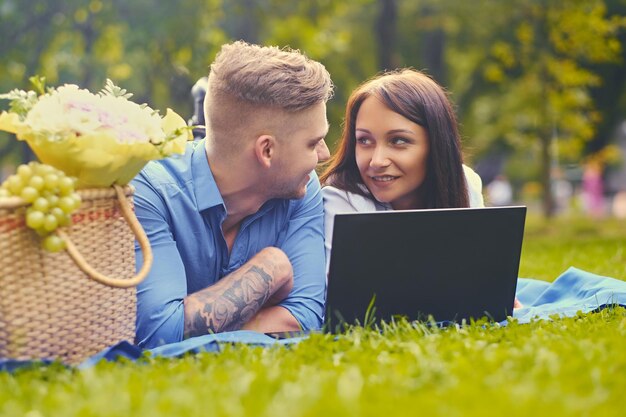 Atrakcyjna para leży na kocu na trawniku i korzysta z laptopa na pikniku.