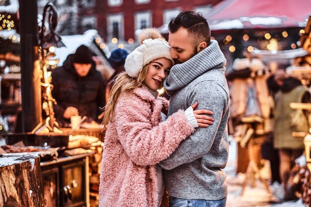 Atrakcyjna para zakochanych, przystojny mężczyzna i urocza dziewczyna przytulająca się i ciesząca się wspólnym spędzaniem czasu, stojąc na zimowym jarmarku w okresie świątecznym