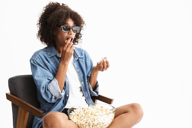 Atrakcyjna podekscytowana młoda kobieta ubrana w dżins, siedząca na krześle na białym tle nad białą ścianą, oglądająca film i jedząca popcorn