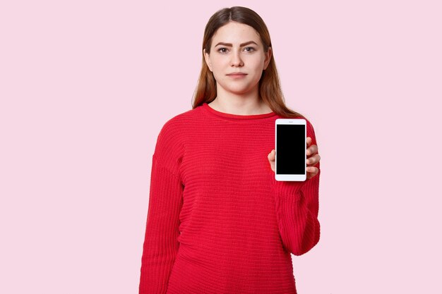 Atrakcyjna poważna młoda europejska nastolatka z długimi włosami, ubrana w czerwony luźny sweter, trzyma nowoczesny telefon komórkowy z czarnym pustym