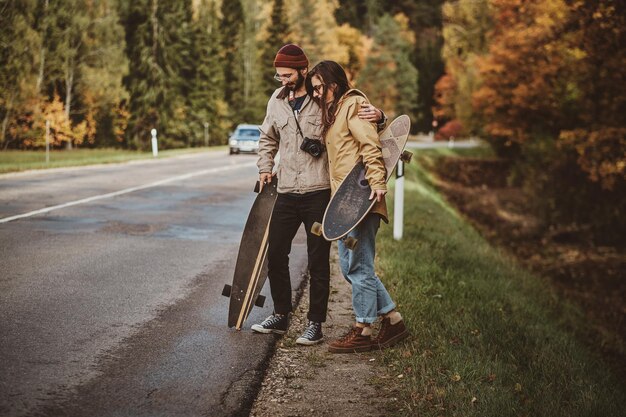 Atrakcyjna romantyczna para idzie drogą otoczoną jesiennymi drzewami trzymając swoje longboardy.