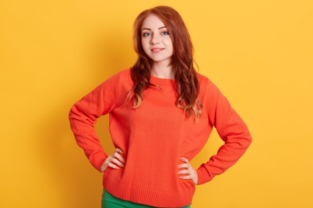 Atrakcyjna rudowłosa dziewczyna patrząc prosto w kamerę z uroczym uśmiechem, ubrana w swobodny pomarańczowy sweter, stojąca przed żółtą ścianą. wyrażanie pozytywnych emocji.