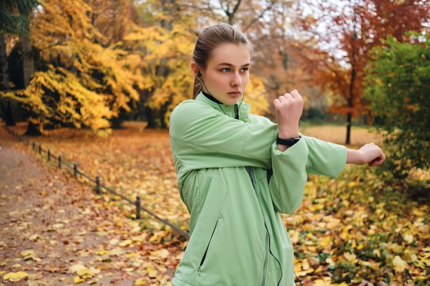 Atrakcyjna sportowa dziewczyna w słuchawkach intensywnie rozciągająca się przed biegiem w jesiennym parku miejskim