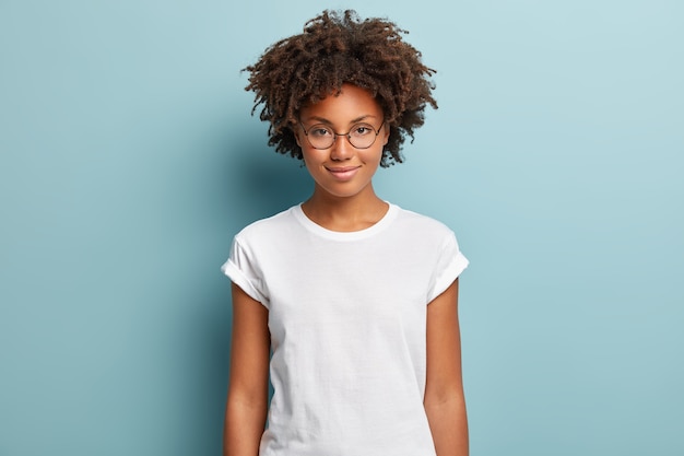 Bezpłatne zdjęcie atrakcyjna studentka o kręconych włosach, nosi przezroczyste okulary, białą koszulkę, stoi na niebieskim tle, ma spokojny wyraz twarzy, delikatny uśmiech,