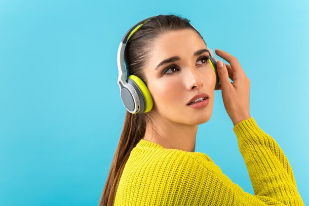 Atrakcyjna, stylowa, młoda kobieta, słuchanie muzyki w słuchawkach bezprzewodowych, szczęśliwy na sobie żółty sweter z dzianiny, kolorowy styl moda pozowanie na białym tle na niebieskiej ścianie