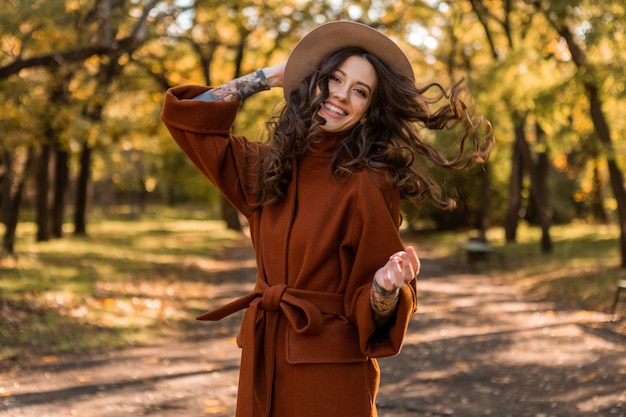 Atrakcyjna stylowa uśmiechnięta chuda kobieta z kręconymi włosami spacerująca po parku ubrana w ciepły brązowy płaszcz, jesień modny styl ulicy