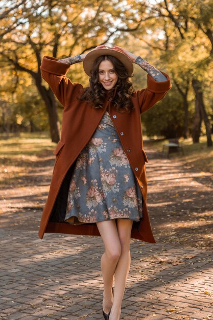 Atrakcyjna, stylowa uśmiechnięta kobieta z kręconymi włosami spacerująca po parku ubrana w drukowaną sukienkę i ciepły płaszcz jesień modna moda, styl uliczny