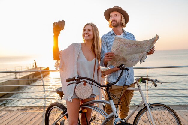 Atrakcyjna szczęśliwa para podróżująca latem na rowerach, mężczyzna i kobieta o blond włosach moda w stylu boho hipster, dobra zabawa razem, patrząc na mapę zwiedzanie, robienie zdjęć aparatem