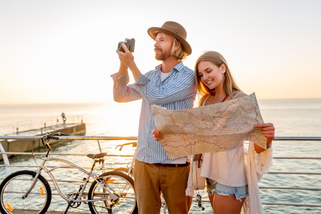 Atrakcyjna szczęśliwa para podróżująca latem na rowerach, mężczyzna i kobieta o blond włosach moda w stylu boho hipster, dobra zabawa razem, patrząc na mapę zwiedzanie, robienie zdjęć aparatem